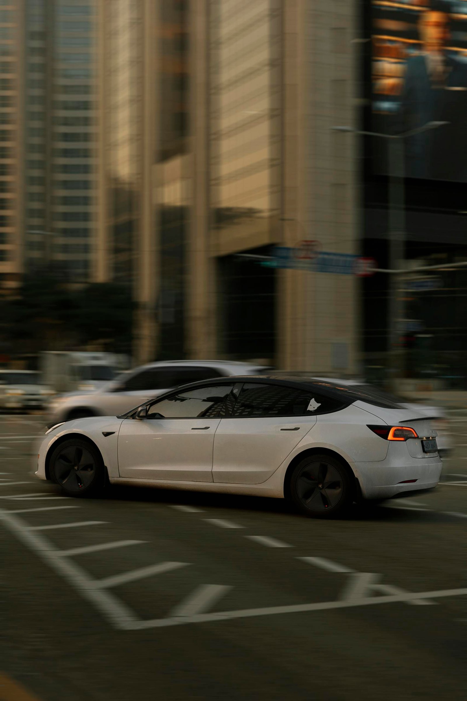 White Tesla Model 3 driving through a busy city intersection with tall buildings.